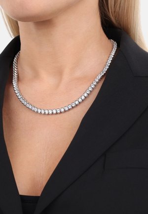 Collana in tonalità argento con cristalli trasparenti rotondi incastonati in una linea continua, poggiante sulla clavicola, indossata con un blazer nero.