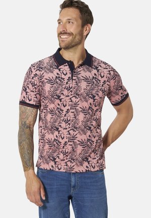 Man with beard smiling, wearing pink floral short-sleeve polo shirt with black collar and blue jeans, standing against plain background.