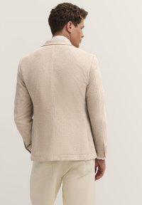 Beige strukturierter Blazer mit einem feinen Gewebe, einem einzelnen Rückenbelüftung und einem Reverskragen. Kombiniert mit hellen Hosen, die einen lässigen Look zeigen.