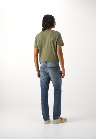 T-shirt in cotone verde oliva con maniche corte, abbinato a jeans in denim blu con due tasche posteriori e lavaggio chiaro. Sneakers bianche con accenti gialli.