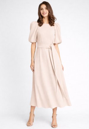 Kobieta ubrana w jasnoróżową sukienkę midi z bufkami przy krótkich rękawach, paskiem wiązanym w talii oraz beżowe wysokie obcasy z paskiem wokół kostki, stojąca na białym tle.