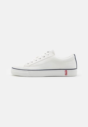 Fehér vászon Sneakers, alacsony profilú dizájnnal, tengerészkék részletekkel a saroknál és a talpnál, valamint hat fűzőlyukkal. Piros logó az oldalon.