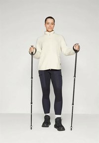 Giacca in pile con zip crema, leggings blu navy con tasca, stivali da trekking neri, in mano bastoni da escursionismo, in piedi su uno sfondo neutro.