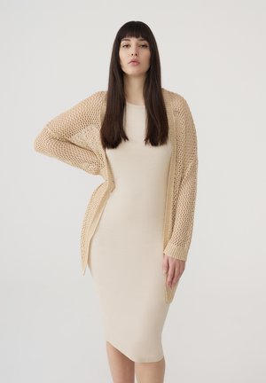 Cardigan beige a maglia con apertura frontale, indossato sopra un vestito aderente beige, che mette in mostra una texture leggera e una silhouette morbida.