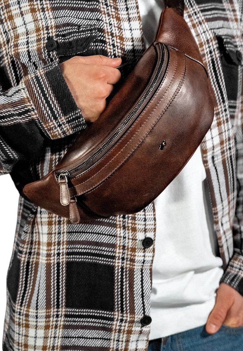 Marsupio in pelle marrone con design curvo, scomparti con doppia zip e un piccolo logo, indossato da una persona con una camicia a quadri.