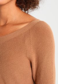 Hnědý pletený svetr s širokým výstřihem a strukturovaným žebrováním, který zdůrazňuje měkkost a pružnost látky.