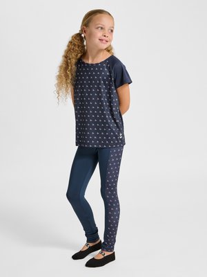 Jong meisje met krullend haar, draagt een marineblauw T-shirt met patroon en legging, staat met handen achter de rug, kijkt opzij en glimlacht.