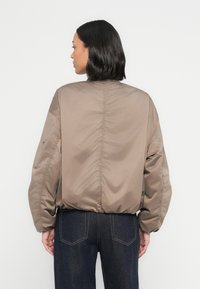 Καφέ μπουφάν τύπου μπόμπερ με υφή επένδυσης, ελαστικά μανίκια και ελαφρώς σούρα πίσω πλευρά. Φοριέται πάνω από σκούρο τζιν, με τσέπη στο μανίκι.