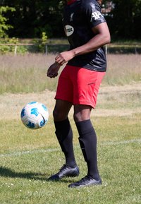 Musta lyhythihainen pelipaita, jossa logo ja punaiset korostukset, punaiset shortsit, mustat sukat ja mustat nappikengät, taitavasti kuljettaa sinivalkoista jalkapalloa.