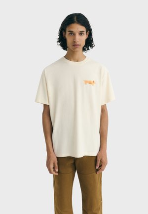 Jong persoon met krullend haar draagt een crèmekleurig T-shirt met een kleine oranje teckel op de borst en een paarse broek, staand voor een effen achtergrond.