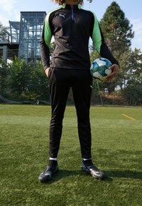 Svart träningsoutfit för sport med långa ärmar och gröna detaljer, tillsammans med svarta fotbollsskor. Personen håller i en flerfärgad fotboll.