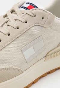 Baskets Tommy Jeans beiges en tissu texturé, détails en daim, lacets, patch logo de la marque, et détail drapeau rouge, blanc et bleu marine sur la languette.
