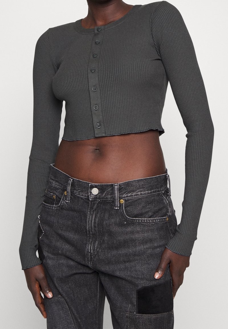 Mörkgrå ribbad kort topp med långa ärmar, med rund halsringning och knapplist fram. Bärs med svarta jeans som visar denimstruktur.