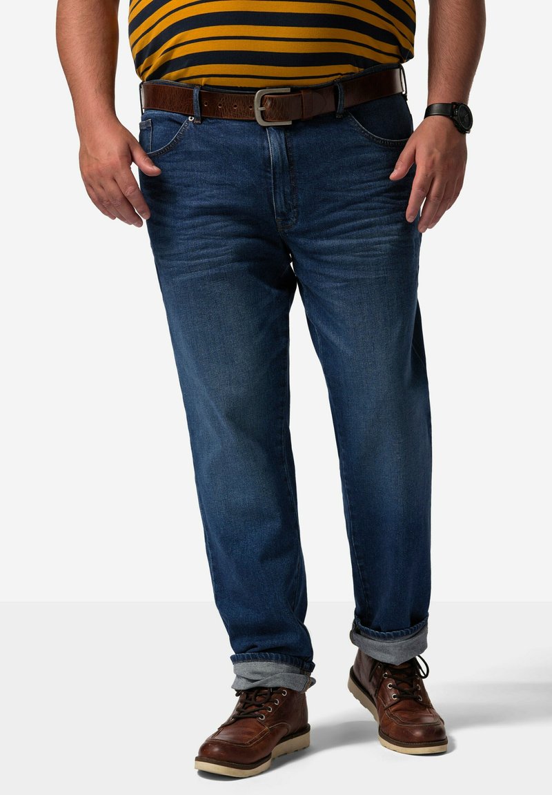 Jeans de denim azul con corte recto, que presentan un desgaste ligero, un cinturón de cuero marrón y los puños enrollados. Llevados con botas de cordones marrones.