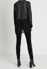 Černý cardigan s dlouhým rukávem z texturované průsvitné látky s vertikálními pruhy, vrstvený přes černé úzké kalhoty a boty na podpatku.