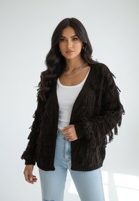 Ciemnobrązowy dziergany kardigan z frędzlami, otwarty z przodu, z prążkowanymi mankietami. Noszony na białej bluzce, w zestawie z jasnoniebieskimi dżinsami.