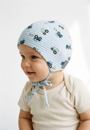 Toddler smiling, wearing a blue striped bonnet with tractor prints, tied under the chin, and a beige short-sleeve shirt.