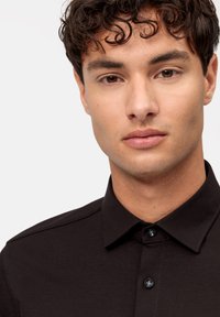 Camicia nera con colletto, caratterizzata da dettagli sulle spalle e due bottoni neri davanti. Tessuto liscio, design aderente.