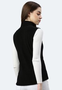 Schwarze ärmellose Weste mit hohem Kragen, kombiniert mit einem weißen gerippten Langarmshirt. Glatte Textur, minimalistisches Design, keine sichtbaren Beschläge oder Muster.