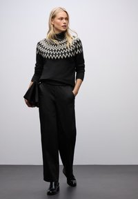 Czarny sweter z golfem z białym wzorem w zygzaki na dekolcie, w połączeniu z luźnymi czarnymi spodniami i czarnymi botkami za kostkę, trzymając czarną kopertówkę.