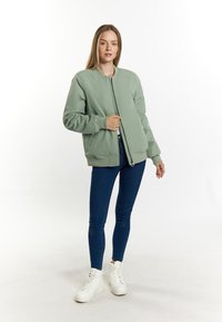 Lichtgroene bomberjack met een ritssluiting aan de voorkant, geribbelde manchetten en onderkant. Gecombineerd met donkerblauwe jeans en witte hoge sneakers.