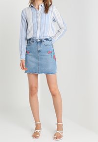 Camicia a righe blu abbinata a una gonna in denim azzurro chiaro con ricami floreali sul davanti. Sandali bianchi con cinturini completano il look.
