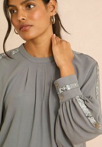 Šedá blůza s plizé a flitrovými pruhy na ramenou a manžetách. Má dlouhé nafouknuté rukávy a kulatý výstřih s ozdobnými knoflíky.