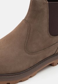 Botină Chelsea din suede maro, cu panou elastic pe laturi, suprafață texturată, talpă din cauciuc contrastantă și detalii de cusătură vizibile.