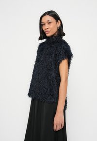 Tmavě modrý chlupatý top bez rukávů s vysokým límcem a texturovaným povrchem, doplněný černou sukní. Top má volný střih a neupravené okraje.