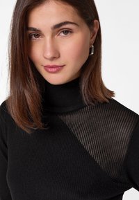 Top noir à col roulé côtelé avec un panneau diagonal transparent sur un côté, mettant en avant un mélange de textures et un design sophistiqué.