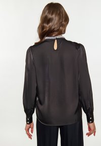 Blusa nera in satin con maniche lunghe, polsini abbottonati e un'apertura a forma di chiave sul retro. Presenta un colletto decorativo con perline.