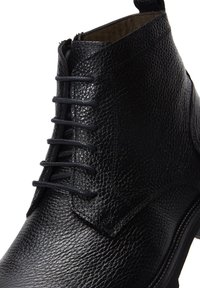 Stivale alla caviglia in pelle nera con una texture a granuli, caratterizzato da un design con lacci, punta arrotondata e dettagli cuciti lungo la suola.