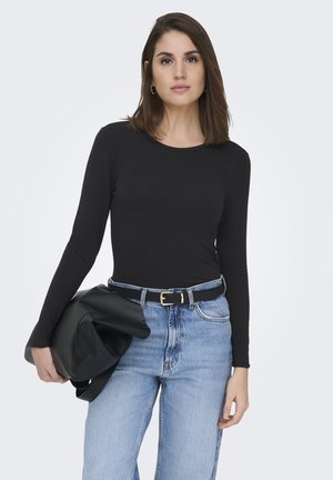 Mujer con cabello castaño hasta los hombros, vestida con una camiseta negra de manga larga, jeans azul claro y sosteniendo una chaqueta negra doblada.