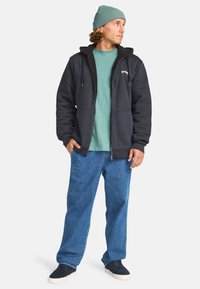 Navy zip-up hoodie with a sherpa-lined hood, paired with a teal shirt and light blue denim jeans. Model wears dark sneakers.