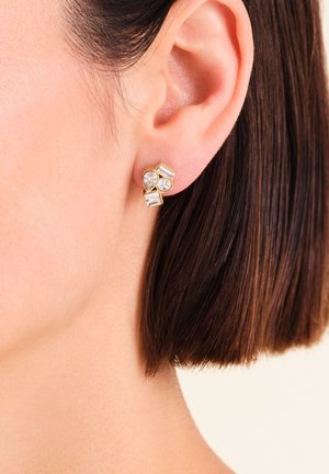 L'oreille d'une femme portant un boucle d'oreille en ton or avec quatre cristaux géométriques transparents en formes de poire, ronde, rectangulaire et baguette.
