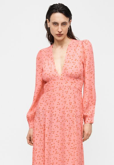 Γυναίκα με σκούρα μαλλιά φοράει ένα μακρυμάνικο ροζ φόρεμα με floral μοτίβο και βαθύ V λαιμόκοψη, στέκεται μπροστά σε ένα απλό λευκό φόντο.