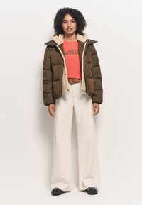 Kurtka puchowa w oliwkowym kolorze z kołnierzem wyłożonym polarem, noszona na koralowej bluzce i beżowych spodniach o wysokim stanie, zestawiona z czarnymi butami.