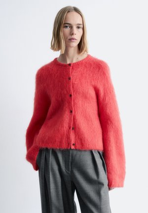 Vrouw met steil blond haar die een zacht koraalkleurig vest met knopen draagt en losse grijze geplooide broek, tegen een effen achtergrond.