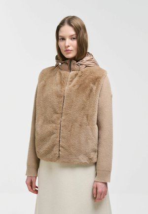 Giovane donna che indossa un gilet in pelliccia sintetica beige con cappuccio sopra un maglione beige lavorato a maglia e una gonna color crema, in piedi davanti a uno sfondo bianco uniforme.