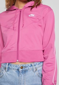 Rosa kort hoodie i slät tyg med dragkedja, med en vit Nike-logotyp och kontrasterande vita ränder längs ärmarna.