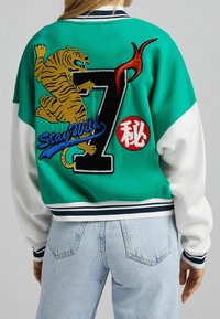 Giacca bomber verde e bianca con un tigre ricamato, il numero '7' e la scritta "Stay Wild" in blu. Polsini e orlo a coste.