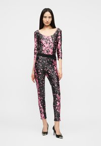 Γυναίκα που στέκεται, φορώντας εφαρμοστό ολόσωμο φόρεμα με ροζ και μαύρο μοτίβο, με μανίκια τριών τετάρτων και μαύρες ψηλοτάκουνες γόβες.