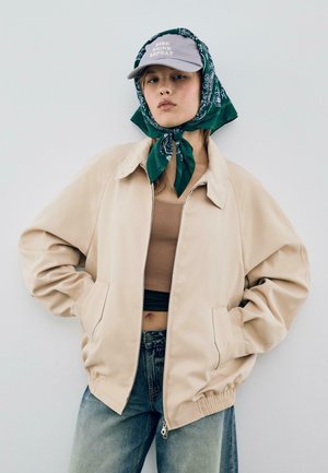 Beige jakke med lynlås og sidelommer, båret over en brun ribbet cropped top. Grøn bandana bundet under en grå kasket med tekst. Denim jeans.