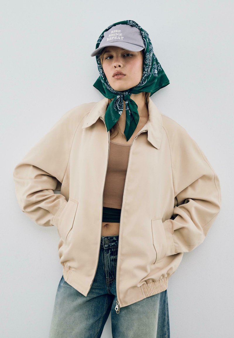 Beige jacka med dragkedja och sidofickor, bärs över en brun ribbad crop top. Grön bandana knuten under en grå keps med text. Denimjeans.