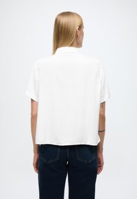 Rövid ujjú, fehér ing enyhén bő szabással és gallérral. Simább textúrájú, egyenes szegéllyel rendelkezik, sötétkék farmerral viselve.