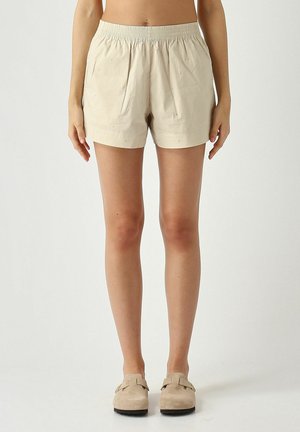 Pantaloncini beige con vita elastica e sottile motivo a piccoli pois, indossati con scarpe slip-on marrone chiaro su sfondo uniforme.