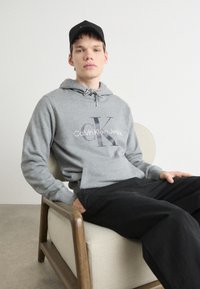 Hoodie grigio in cotone con cappuccio e tasca anteriore, caratterizzato da un logo nero. Abbinato a pantaloni neri, modello seduto su una sedia chiara.