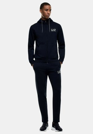 Giovane uomo in piedi con le mani nelle tasche, indossa una felpa nera EA7 Emporio Armani con zip e pantaloni da jogging abbinati con sneakers nere.