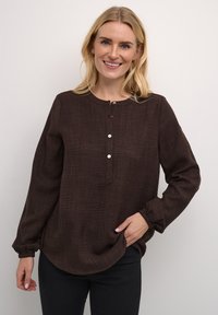 Haut long à manches marron avec un col rond, fermeture à boutons sur le devant et coupe décontractée. Associé à un pantalon noir.