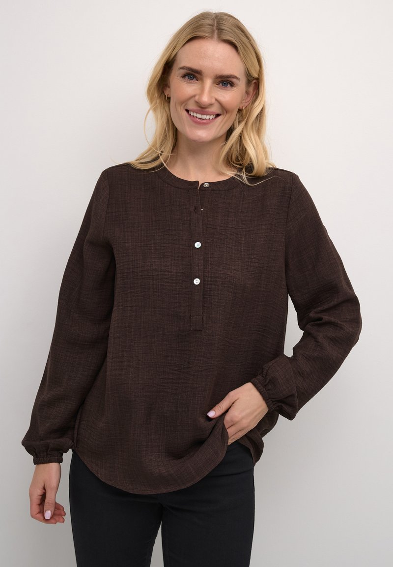 Haut long à manches marron avec un col rond, fermeture à boutons sur le devant et coupe décontractée. Associé à un pantalon noir.
